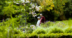 An elderly couple walking hand in hand through a lush, vibrant garden filled with blooming flowers, rich greenery, and hints of autumn colors. This image embodies the theme of The Healing Power of Nature: A Holistic Exploration, symbolizing the joy, calm, and rejuvenating effect of being immersed in natural surroundings, as well as the healing presence of shared moments in a tranquil setting.