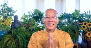 Master Mingtong Gu is smiling warmly with his hands in a prayerful gesture, dressed in a golden, embroidered traditional outfit. He is surrounded by lush greenery, vibrant flowers, and a serene Buddha statue in the background, creating a peaceful and harmonious atmosphere. The image exudes warmth, wisdom, and a sense of calm energy.