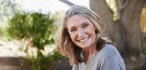 Middle-aged woman with a warm, joyful smile, outdoors in natural sunlight, surrounded by greenery, embodying health, vitality, and contentment, symbolizing the benefits of Wisdom Healing Qigong for health and healing.