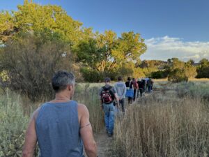 A group of people walk in single file along a narrow trail through tall grasses and sagebrush, heading toward a large, majestic tree known as the 'Grandmother Tree.' The sun casts a warm glow on the landscape, highlighting the natural beauty of the scene. The journey appears peaceful and contemplative, symbolizing a shared connection to nature and the wisdom embodied by the Grandmother Tree.