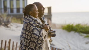 An older couple stands on a sandy beach, wrapped together in a cozy, patterned blanket, each holding a mug. They smile warmly as they look out toward the ocean, enjoying a peaceful moment at sunrise or sunset. The relaxed setting and their joyful expressions capture a sense of togetherness, warmth, and appreciation for life’s simple pleasures.