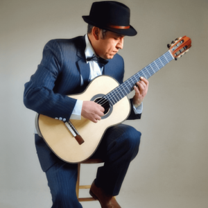 Tito Rios, dressed in a dark suit, white shirt, and black bow tie, sits on a stool playing a classical guitar with focused intensity. He wears a black fedora hat, his gaze directed at the guitar strings as he skillfully performs.