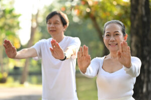 An older couple stands on a sandy beach, wrapped together in a cozy, patterned blanket, each holding a mug. They smile warmly as they look out toward the ocean, enjoying a peaceful moment at sunrise or sunset. The relaxed setting and their joyful expressions capture a sense of togetherness, warmth, and appreciation for life’s simple pleasures.