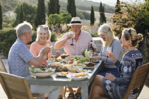 A group of older adults enjoying a cheerful outdoor gathering around a table laden with food and drinks. They are seated in a sunny, scenic setting with a view of trees and hills in the background. Smiling and raising their glasses in a toast, they share a variety of fresh fruits, vegetables, bread, and cheeses, embodying a sense of community, relaxation, and joy in each other’s company.