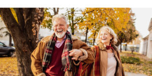 A joyful older couple walks arm in arm on a crisp autumn day, laughing and enjoying each other's company. Dressed warmly in cozy coats and colorful scarves, they stroll through a park filled with vibrant fall foliage. Their radiant smiles and lively energy capture a sense of vitality, companionship, and appreciation for life, embodying the spirit of embracing one’s golden years