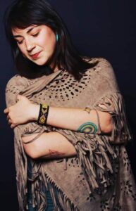 "Speaker for Magical Sundays wraps herself in a fringed shawl, looking down peacefully, wearing a colorful beaded bracelet and arm tattoos."