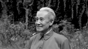 Portrait of Dr. Pang, a revered figure in Qigong practice, smiling and looking thoughtfully ahead, set against a natural, leafy backdrop.