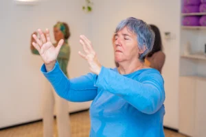 Woman practicing Wisdom Healing Qigong in a calm indoor space, focusing on gentle hand movements and energy flow, with others practicing in the background.