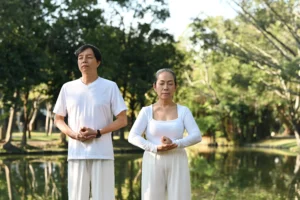 Man and woman practicing Qigong by a serene lake, eyes closed and hands in a mudra, as part of a 7 Weeks of Sound Healing focus and practice.