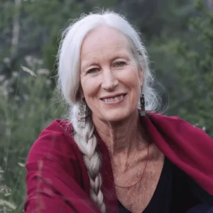 This image shows Cynthia Jurs, she has long white hair braided over her shoulder, wearing a deep red shawl. She has a warm and gentle smile, and her surroundings are filled with greenery, giving a sense of being in nature. Her earrings and subtle jewelry add an earthy, spiritual touch, reflecting a peaceful and wise presence.