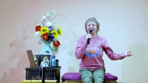Kira Schofield enthusiastically shares testimonials during a fall retreat, holding a microphone and gesturing warmly. Dressed in a vibrant sweater, she sits beside a colorful floral arrangement and spiritual icons, creating an inviting atmosphere filled with openness, connection, and community healing.