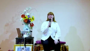 Lisa shares heartfelt testimonials at a fall retreat, seated with a microphone in hand and a warm, reflective expression. Dressed in a light blouse, she sits beside a vibrant floral arrangement and spiritual icons, creating a welcoming atmosphere for community connection and personal growth.