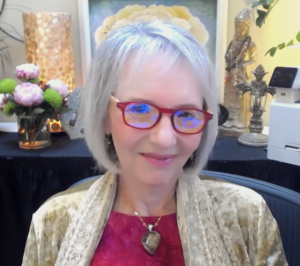 This image shows Sallena Pool, a woman with short, silver hair and red-rimmed glasses, smiling warmly. She is dressed in a golden-beige shawl over a red top, with a heart-shaped pendant necklace. The serene background features flowers, a lit lamp, a Buddha statue, and various decorative artifacts, creating a peaceful, spiritual atmosphere. Sallena Pool appears calm and relaxed in this harmonious setting.