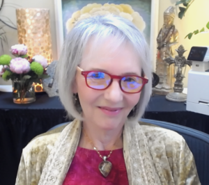 This image shows Sallena Pool, a woman with short, silver hair and red-rimmed glasses, smiling warmly. She is dressed in a golden-beige shawl over a red top, with a heart-shaped pendant necklace. The serene background features flowers, a lit lamp, a Buddha statue, and various decorative artifacts, creating a peaceful, spiritual atmosphere. Sallena Pool appears calm and relaxed in this harmonious setting.