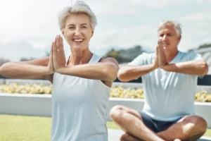 An elderly woman and man practicing mindful meditation outdoors, sitting cross-legged and holding their hands in a prayer position. They smile peacefully, embodying health, vitality, and the joy of wellness practices in a serene natural setting.