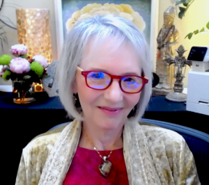 This image shows Sallena Pool, a woman with short, silver hair and red-rimmed glasses, smiling warmly. She is dressed in a golden-beige shawl over a red top, with a heart-shaped pendant necklace. The serene background features flowers, a lit lamp, a Buddha statue, and various decorative artifacts, creating a peaceful, spiritual atmosphere. Sallena Pool appears calm and relaxed in this harmonious setting.