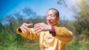 Master Mingtong Gu practices Qigong outdoors, wearing a golden traditional robe that glows in the sunlight. His eyes are closed, and his hands are held in a graceful, focused position, embodying tranquility and inner connection. The natural green backdrop and clear blue sky add to the serene, healing atmosphere. This image captures the essence of Qigong for Healing, symbolizing the fusion of ancient wisdom with modern approaches to wellness and holistic medicine.
