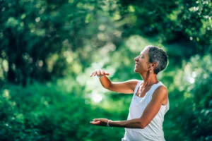 An image of a woman practicing Qigong outdoors, surrounded by lush green foliage, exuding a sense of peace and well-being. She is smiling gently, with her hands raised in a graceful posture, embodying the balance of energy flow. This serene image symbolizes the harmony between modern medicine and Qigong, showcasing how ancient energy practices can support holistic health and healing in a natural setting