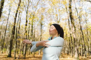 A young woman practices Qigong in a serene forest, surrounded by tall trees with autumn leaves. She stands with her arms crossed in a gentle stretch, eyes closed, and a peaceful expression on her face, embodying calmness and connection to nature. This image reflects the integration of ancient wisdom with modern practices for healing, symbolizing grounding, immunity, and holistic well-being through Qigong in a natural environment.