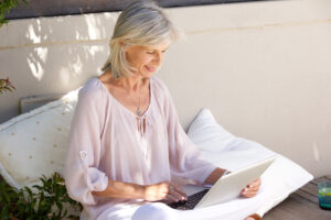 Elderly woman with silver hair working on a laptop outdoors, sitting on a cozy patio with soft cushions and natural light. Perfect for themes of relaxation, remote work, or staying connected in a serene environment.
