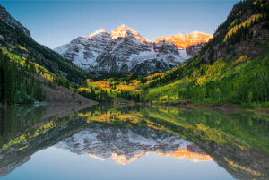 Snow-dusted mountain peaks glowing with golden sunlight under a clear blue sky, surrounded by lush green valleys and forested slopes. A perfect depiction of natural beauty and serenity in the great outdoors.