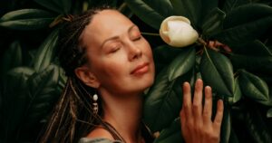 A serene woman with braided hair gently embraces a blooming white flower amidst lush green leaves, her eyes closed in peaceful contemplation. The image reflects a deep connection with nature and serving from an awakened consciousness.