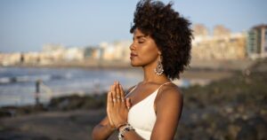 A serene woman standing on a beach at sunset, hands in prayer position near her heart, embodying tranquility and intention. Her calm expression and graceful posture reflect serving from an awakened consciousness, with a backdrop of waves and a distant cityscape.