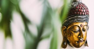A close-up of a golden Buddha statue with intricate details and a serene expression, partially obscured by soft green leaves in the foreground.