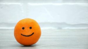 An orange with a cute smiley face drawn on it, sitting on a light wooden surface against a white brick wall background. Perfect for themes of positivity, healthy living, and playful creativity.