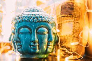 A vibrant turquoise Buddha head sculpture with multiple serene faces, surrounded by warm string lights and a golden textured cushion in the background. Perfect for themes of mindfulness, spirituality, and peaceful decor.