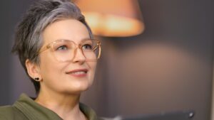 A confident woman with short gray hair and large glasses smiles softly while looking upward, sitting indoors with warm lighting in the background.