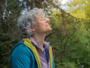 A joyful woman with gray curls enjoys a peaceful moment outdoors, closing her eyes and smiling while taking a deep breath in a forested area, symbolizing relaxation, connection with nature, and mindfulness.
