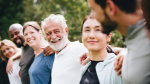 A diverse group of people standing outdoors in a line with their arms around each other's shoulders, smiling warmly and looking happy in a natural setting.