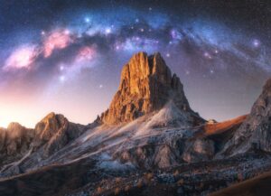 A stunning mountain peak illuminated by soft sunlight, surrounded by rocky slopes and a star-filled sky featuring the vibrant arc of the Milky Way.