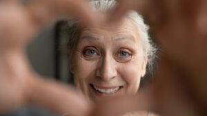 A cheerful older woman with gray hair smiles warmly while framing her face with her hands, creating a heart shape in the foreground.