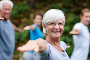 A joyful senior woman with white hair practicing outdoor group yoga or fitness, smiling confidently at the camera while stretching her arms forward. A diverse group of individuals participates in the background.
