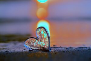 A transparent heart-shaped glass ornament rests on a wet surface, reflecting the warm hues of a glowing sunset or sunrise in the background, creating a serene and romantic atmosphere.