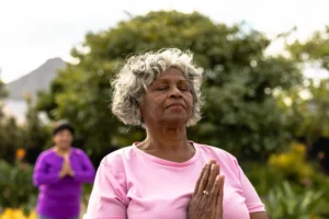 An older woman with curly gray hair wearing a pink shirt stands outdoors with eyes closed and hands in a prayer position at her chest, radiating calm and focus.