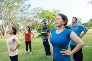 A diverse group of adults practicing Qigong outdoors in a lush green park, standing with hands on hips, eyes closed, and deep breathing, embracing relaxation, energy flow, and mindful movement for health and well-being.