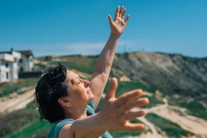 A woman with short dark hair stands outdoors on a sunny day with her eyes closed and arms joyfully raised toward the sky.