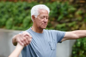 An older man with white hair wearing a light blue t-shirt is practicing a meditative movement outdoors.