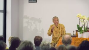 Master Mingtong Gu teaching Wisdom Healing Qigong to a live audience, wearing a golden traditional jacket and speaking with expressive hand gestures, seated beside orchids and sacred objects on a table—capturing a moment of healing wisdom and spiritual connection.