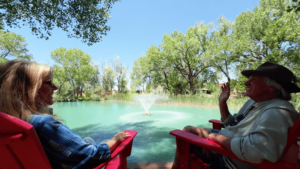 Two people sit in bright red chairs by a turquoise pond with a central fountain, surrounded by tall green trees under a clear blue sky at The Southwest Sanctuary.