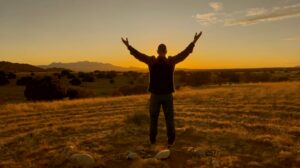 Master Mingtong Gu stands in an open field at sunset with arms uplifted toward the sky, silhouetted by golden light over the New Mexico landscape.