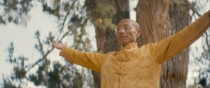 Master Mingtong Gu practicing Qigong outdoors with arms open wide, wearing a golden traditional Chinese jacket, standing peacefully in front of tall trees—symbolizing healing, energy flow, and connection with nature.