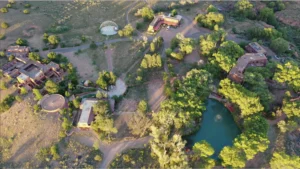 Aerial view of The Southwest Sanctuary retreat center in Galisteo, New Mexico, showcasing adobe-style buildings, walking paths, a circular gathering area, and a serene pond surrounded by trees.