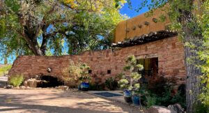 Main lodge at The Southwest Sanctuary in Galisteo, New Mexico, featuring a curved stone façade, natural adobe architecture, and a shaded wooden pergola. Surrounded by vibrant greenery, large cottonwood trees, blue sky, and potted plants, the space reflects harmony with nature and offers a serene setting for retreats, healing, and spiritual awakening.