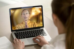 A person sits at a desk using a laptop, watching an online video of Master Mingtong wearing a gold traditional-style robe with hands pressed together in a greeting gesture. Master Mingtong appears on the screen against a warm golden background labeled “Wisdom Healing Qigong.” The viewer’s hands rest on the keyboard, suggesting participation in an online class or guided practice.
