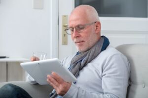An older man with short gray hair and glasses sits on a light-colored sofa, looking down at a tablet he holds in both hands while also holding a glass. He wears a light sweater and a scarf, and the bright, minimalist room behind him gives a calm, comfortable feeling.