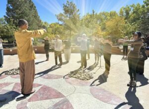 A group of adults stands outdoors in a circular formation, following Master Mingtong in a gold jacket who faces them with arms extended. The participants mirror the movement with their arms held forward at chest height. Sunlight streams through trees in the background, casting warm rays across the scene, which takes place on a patterned stone courtyard surrounded by greenery, creating a peaceful group practice atmosphere.